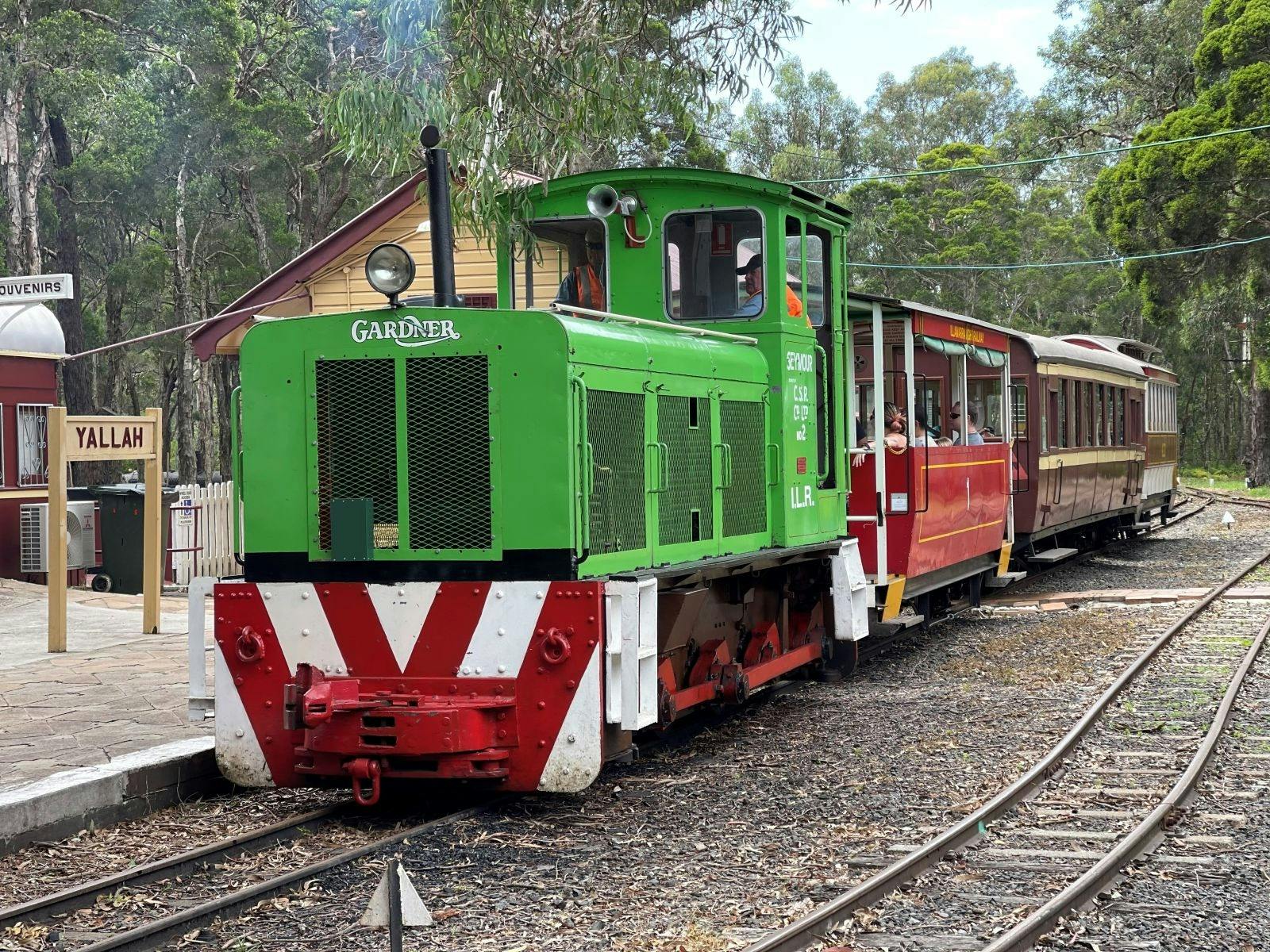 Green train with three brown carriages pulling up to a platform at Illawarra Light Railway Museum